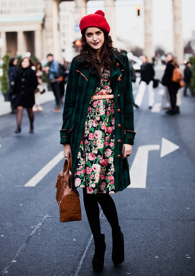 A brunette woman wears a red beret and cute floral dress with a green coat and clashing spring colors